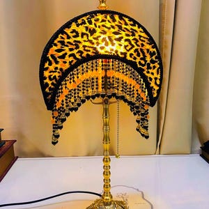 Puede incluir: Lámpara de mesa decorativa con base y tallo dorados. La pantalla presenta un diseño de estampado de leopardo con ribete negro y flecos de cuentas. La lámpara está encendida, emitiendo un brillo cálido.