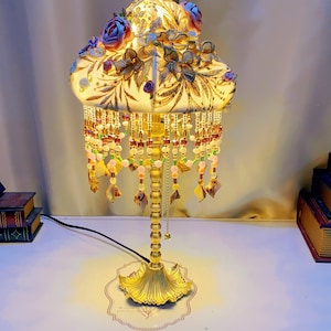 May include: A decorative table lamp with a floral design. The lamp features a cream-colored shade adorned with blue and purple roses and brown floral accents. The base is gold-colored, and the lamp is embellished with hanging beads. The text "Bright freedom" is at the base.