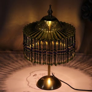 Puede incluir: Una l&aacute;mpara de mesa con una pantalla de vidrio verde oliva plisada y un remate superior puntiagudo. La pantalla est&aacute; adornada con hileras de cuentas negras, moradas y verdes. La l&aacute;mpara tiene un tallo de metal dorado fino y una base circular.