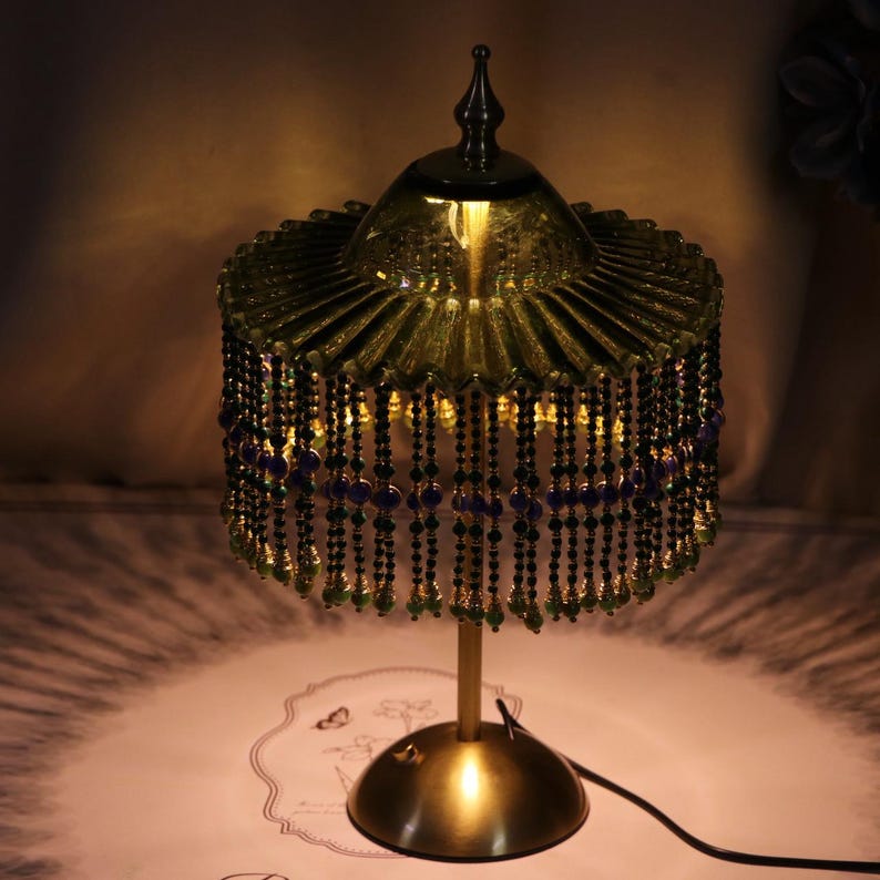 Puede incluir: Una l&aacute;mpara de mesa con una pantalla de vidrio verde oliva plisada y un remate superior puntiagudo. La pantalla est&aacute; adornada con hileras de cuentas negras, moradas y verdes. La l&aacute;mpara tiene un tallo de metal dorado fino y una base circular.