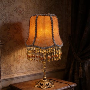 May include: An antique-style table lamp with a golden base and a lampshade in shades of orange and brown. The lampshade is adorned with beaded fringe and decorative trim. The lamp is placed on a wooden table.