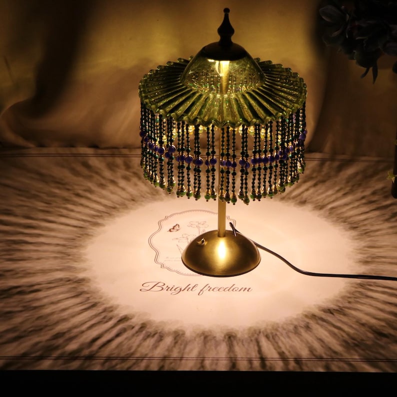 Puede incluir: Una l&aacute;mpara de mesa con base dorada y pantalla de cristal verde plisado con flecos de cuentas colgantes en negro, verde y azul. La l&aacute;mpara proyecta un patr&oacute;n de luz circular sobre la superficie inferior, que muestra el texto Bright freedom.