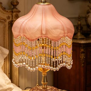 May include: A table lamp featuring a scalloped pink fabric shade with a gold finial. The shade is adorned with hanging strands of pink and yellow beads ending in clear teardrop crystals. The lamp has a gold metal stand and a pink glass base.
