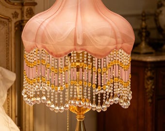 Puede incluir: Una lámpara de mesa con una pantalla de tela rosa festoneada y un remate dorado. La pantalla está adornada con hilos colgantes de cuentas rosas y amarillas que terminan en cristales transparentes en forma de lágrima. Tiene un soporte de metal dorado y una base de cristal rosa.