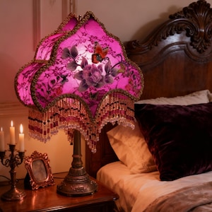 May include: A vintage-style table lamp with a fuchsia shade featuring floral and butterfly designs. The shade is trimmed with gold and beaded fringe. The lamp sits on a decorative bronze base, next to a candle holder and a framed picture.