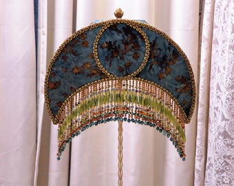 May include: A decorative lamp with a teal and gold floral patterned shade. The shade is trimmed with gold braid and a beaded fringe in shades of green, orange, and teal. The lamp has a gold-colored metal stand.