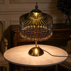 Puede incluir: Una l&aacute;mpara de mesa con base color lat&oacute;n y pantalla de cristal verde transl&uacute;cido plisado. La pantalla est&aacute; adornada con m&uacute;ltiples hileras de cuentas negras y moradas colgantes, cada una con un peque&ntilde;o colgante de cristal verde en forma de gota.