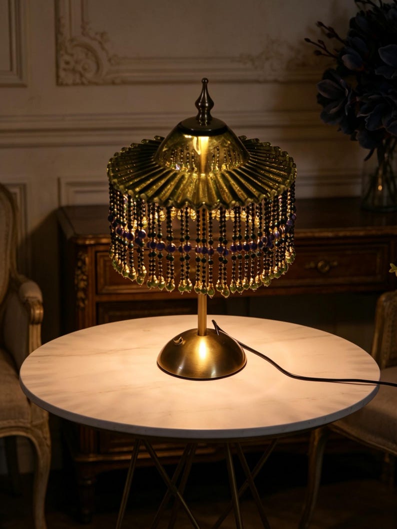 Puede incluir: Una l&aacute;mpara de mesa con base color lat&oacute;n y pantalla de cristal verde transl&uacute;cido plisado. La pantalla est&aacute; adornada con m&uacute;ltiples hileras de cuentas negras y moradas colgantes, cada una con un peque&ntilde;o colgante de cristal verde en forma de gota.