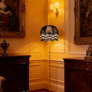 May include: A floor lamp with a black lace shade and a gold-colored metal base and pole. The shade has a floral pattern and is trimmed with white fringe and black beads. The lamp is in a room with antique furniture and artwork.