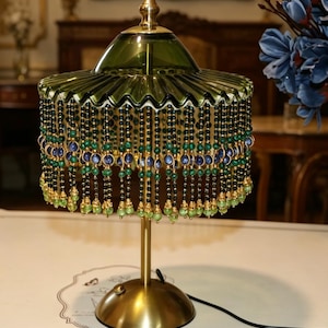 Puede incluir: Una l&aacute;mpara de mesa con base color lat&oacute;n y pantalla de cristal verde con borde plisado. La pantalla est&aacute; adornada con hilos colgantes de cuentas verde oscuro, azules y verde claro. Un peque&ntilde;o interruptor est&aacute; en la base y un cable negro se extiende.