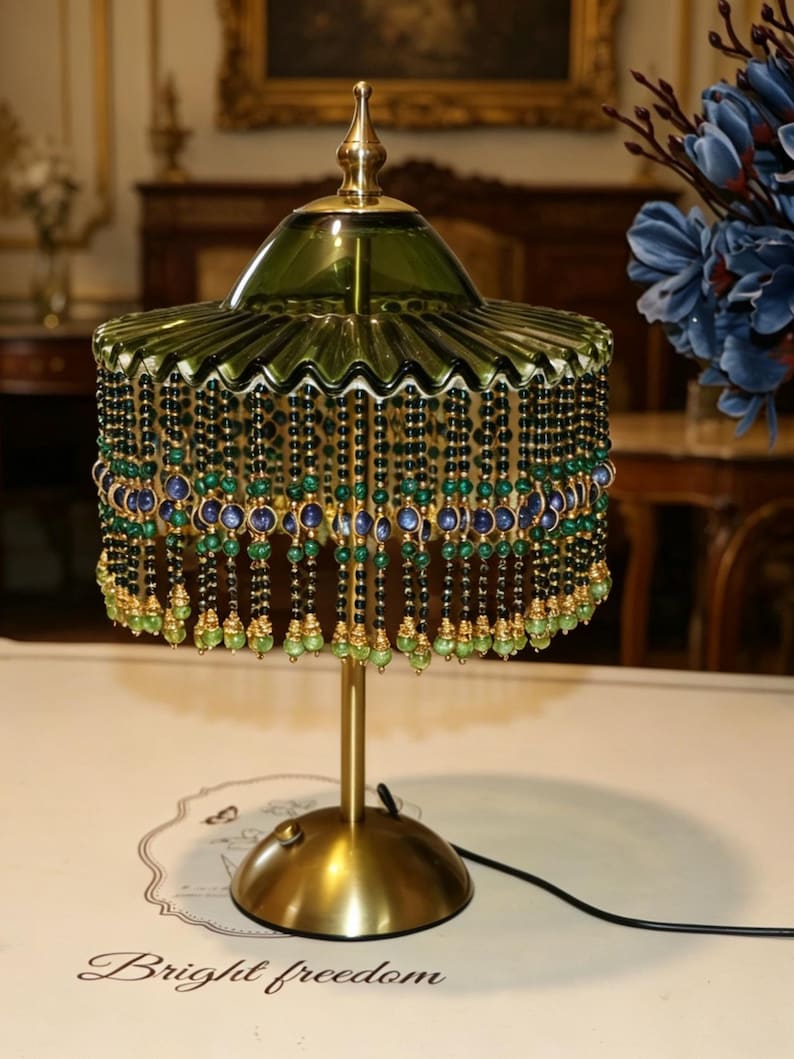 Puede incluir: Una l&aacute;mpara de mesa con base color lat&oacute;n y pantalla de cristal verde con borde plisado. La pantalla est&aacute; adornada con hilos colgantes de cuentas verde oscuro, azules y verde claro. Un peque&ntilde;o interruptor est&aacute; en la base y un cable negro se extiende.