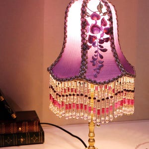 Puede incluir: Una lámpara de mesa morada con flecos de cuentas y bordados florales en la pantalla. La lámpara tiene una base dorada con un diseño ornamentado.
