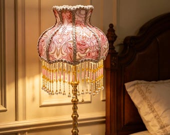 Può includere: Lampada da tavolo in stile vintage con paralume a fantasia floreale rosa e base color oro. Il paralume è decorato con frange di perline. La lampada è posizionata su un comodino in legno, creando un'atmosfera calda. Altezza: circa 50 cm.