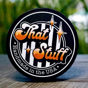 May include: A round black sticker with the words "That Stuff" in orange and white, with a starburst design. The sticker also says "Handmade in the USA" in white text around the edge. The background is blurred.