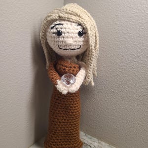 May include: A handmade crochet doll with a light skin tone, blonde hair, and a brown dress. The doll is holding a clear, faceted gem. The doll's face has black eyes and a simple smile. The doll is standing in a corner.