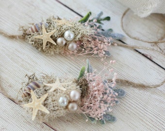 May include: Two delicate floral arrangements with starfish and pearls, tied with twine, on a white wooden background.