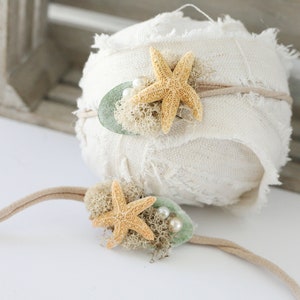 May include: Two beige fabric headbands with starfish and green accents. The headband on the left has two white pearls. The headband on the right has a single white pearl.