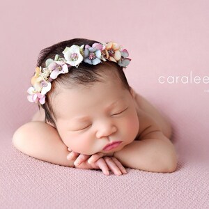 Puede incluir: Una reci&eacute;n nacida con una corona de flores rosas, blancas y azules, durmiendo sobre una manta rosa.