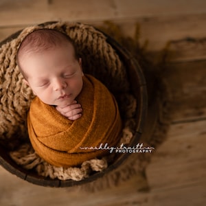 Könnte beinhalten: Ein neugeborenes Baby ist in eine braune Decke gewickelt und schläft in einer Holzschale, die mit beigefarbenem Garn ausgekleidet ist. Die Augen des Babys sind geschlossen und seine Hände sind eingekuschelt. Die Schale steht auf einer Holzoberfläche.