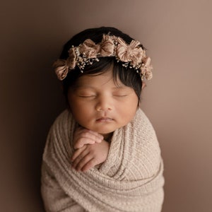 May include: A newborn baby girl is sleeping peacefully, wrapped in a beige blanket. She is wearing a flower crown with brown and white flowers. The background is a soft brown color.