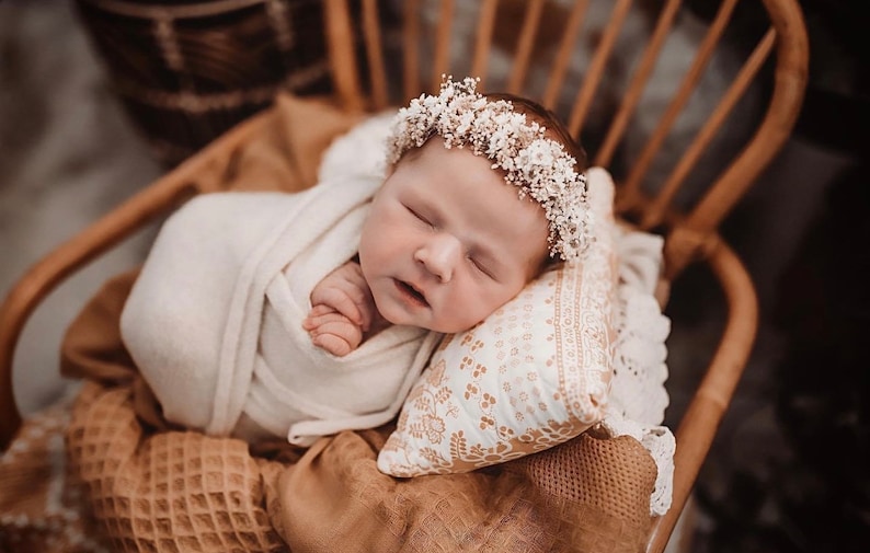 May include: A newborn baby girl wearing a white flower crown sleeps peacefully on a white pillow with a floral pattern. The baby is wrapped in a white blanket and is lying on a brown woven chair.