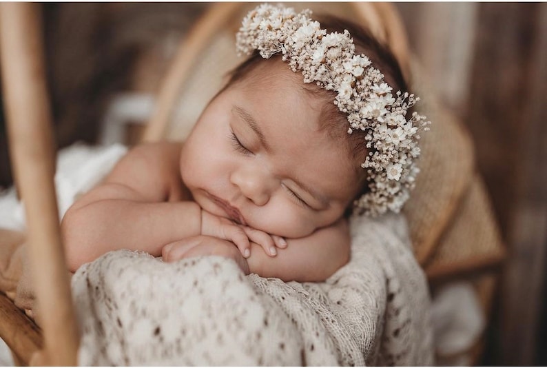 May include: A sleeping baby girl wearing a white flower crown, lying on a white crocheted blanket in a wicker basket.