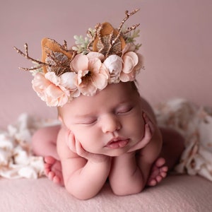 May include: A newborn baby wearing a flower crown with antlers and a brown felt base. The crown has pink and white flowers and green leaves. The baby is sleeping with their hands under their chin.