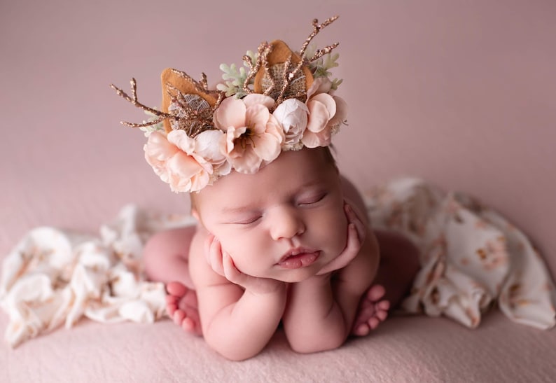 May include: A newborn baby wearing a flower crown with antlers and a brown felt base. The crown has pink and white flowers and green leaves. The baby is sleeping with their hands under their chin.