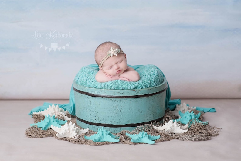 May include: A newborn baby girl sleeping in a turquoise blue wooden bucket with a white starfish headband. The bucket is surrounded by white and blue starfish on a brown burlap surface.