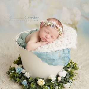 Puede incluir: Un beb&eacute; reci&eacute;n nacido con una corona de flores duerme pl&aacute;cidamente en un cubo blanco con un borde de encaje azul. El cubo est&aacute; rodeado de una guirnalda verde con flores blancas y azules.