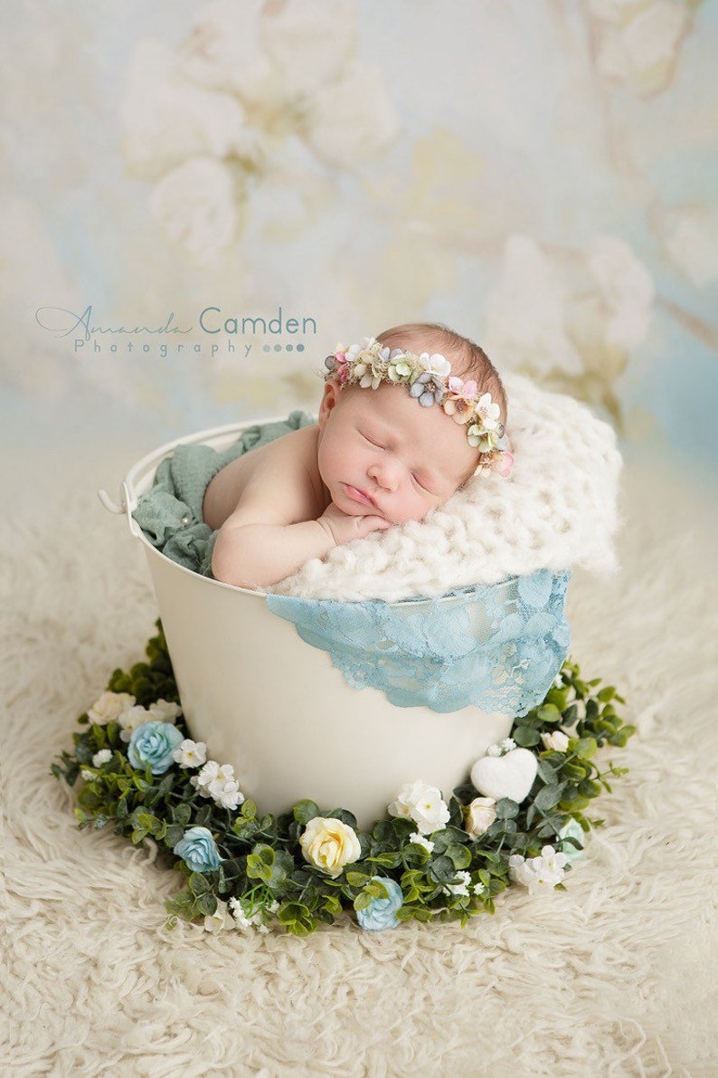 Puede incluir: Un beb&eacute; reci&eacute;n nacido con una corona de flores duerme pl&aacute;cidamente en un cubo blanco con un borde de encaje azul. El cubo est&aacute; rodeado de una guirnalda verde con flores blancas y azules.