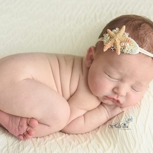 May include: A sleeping newborn baby wearing a headband with a starfish and pearl embellishment. The baby is lying on a white fluffy blanket.
