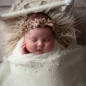 May include: A newborn baby girl is sleeping peacefully in a white swaddle with pearl accents. She is wearing a brown flower headband. The baby is lying on a white furry surface.