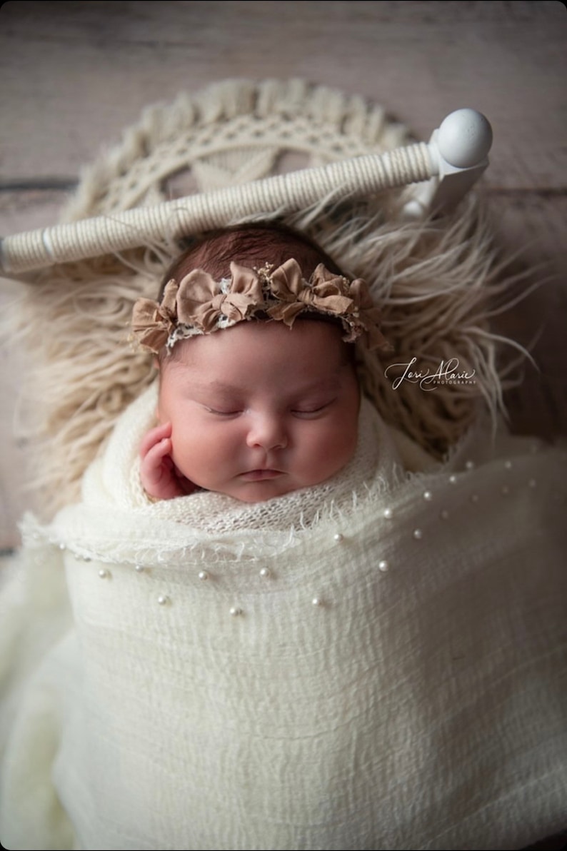May include: A newborn baby girl is sleeping peacefully in a white swaddle with pearl accents. She is wearing a brown flower headband. The baby is lying on a white furry surface.