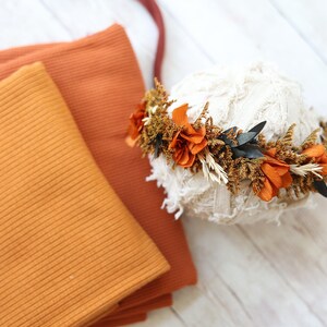 Könnte beinhalten: Zwei orangefarbene gerippte Stoffproben und eine handgefertigte Blumenkrone mit getrockneten orangefarbenen und braunen Blumen. Die Krone liegt auf einer weißen, strukturierten Oberfläche.