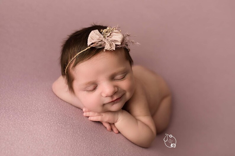 May include: A newborn baby girl is sleeping on a pink blanket. She is wearing a headband with a flower and a bow.