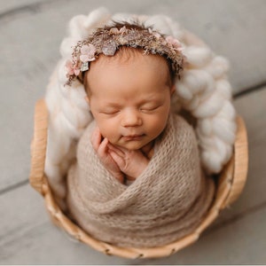 May include: A newborn baby is sleeping peacefully in a woven basket. The baby is wrapped in a beige knitted blanket and wearing a delicate floral headband with a gold accent.