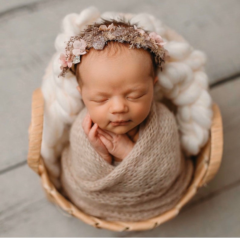 May include: A newborn baby is sleeping peacefully in a woven basket. The baby is wrapped in a beige knitted blanket and wearing a delicate floral headband with a gold accent.