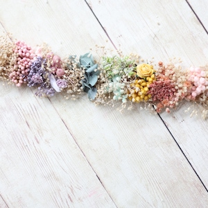 Puede incluir: Una guirnalda de flores secas de colores arco&iacute;ris, incluyendo flores rosas, moradas, azules, verdes, amarillas y naranjas. La guirnalda est&aacute; sobre un fondo de madera blanca.