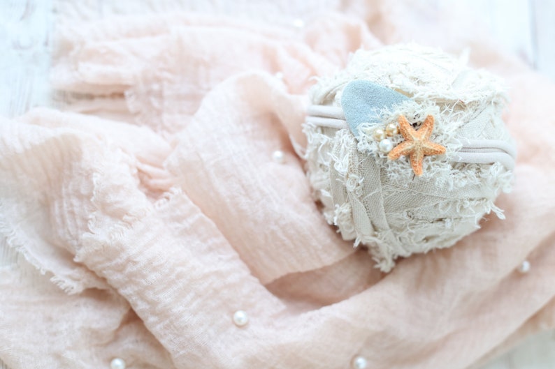 May include: A white fabric headband with a light blue felt starfish and a small orange starfish. The headband is on a pink fabric background.
