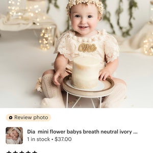 May include: A baby girl wearing a white lace dress and a floral headband with the word "One" on her chest, sitting in front of a white cake on a wooden cake stand.