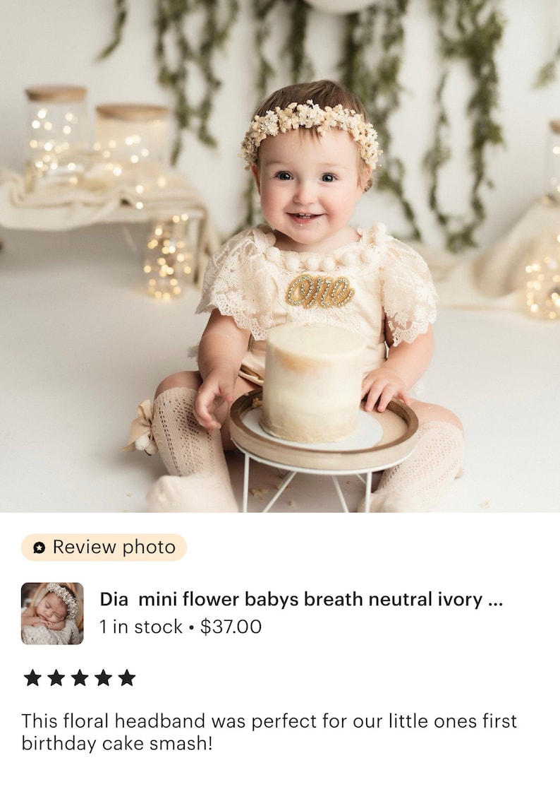 May include: A baby girl wearing a white lace dress and a floral headband with the word "One" on her chest, sitting in front of a white cake on a wooden cake stand.