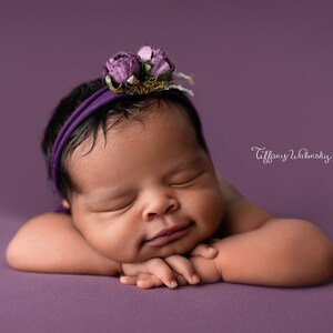 Peut inclure: Un nouveau-né dort sur un fond violet. Elle porte un bandeau violet avec une fleur sur le dessus. Le bébé repose sa tête sur ses bras et ses yeux sont fermés.