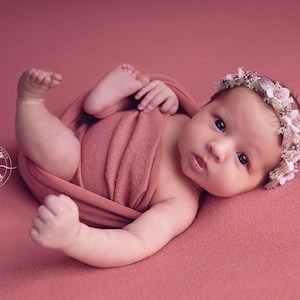 May include: A newborn baby girl wearing a pink flower crown, wrapped in a peach-colored swaddle, lying on a pink background.