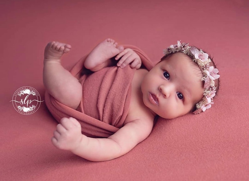 May include: A newborn baby girl wearing a pink flower crown, wrapped in a peach-colored swaddle, lying on a pink background.