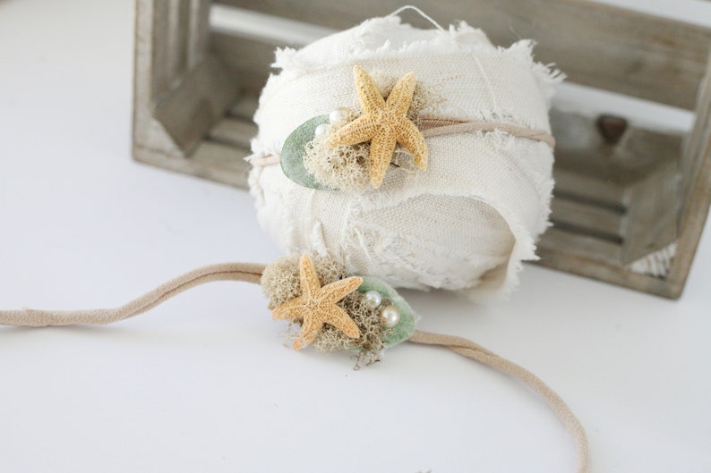 May include: Two beige fabric headbands with starfish and green accents. The headband on the left has two white pearls. The headband on the right has a single white pearl.