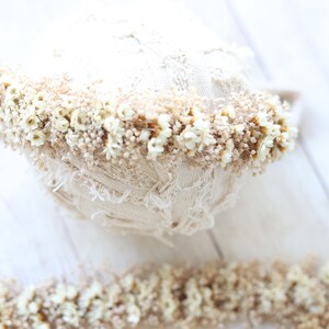 May include: A white lace headband with a band of dried white flowers and tan stems. The headband is on a white wooden surface.