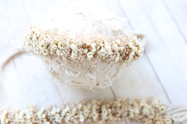 May include: A white lace headband with a band of dried white flowers and tan stems. The headband is on a white wooden surface.