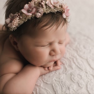 May include: A newborn baby girl wearing a pink flower crown, sleeping on a white textured blanket.