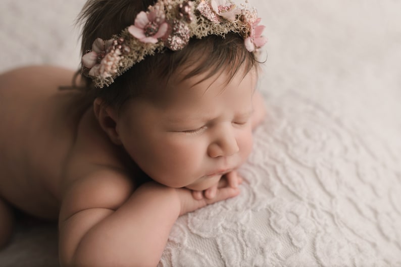 May include: A newborn baby girl wearing a pink flower crown, sleeping on a white textured blanket.